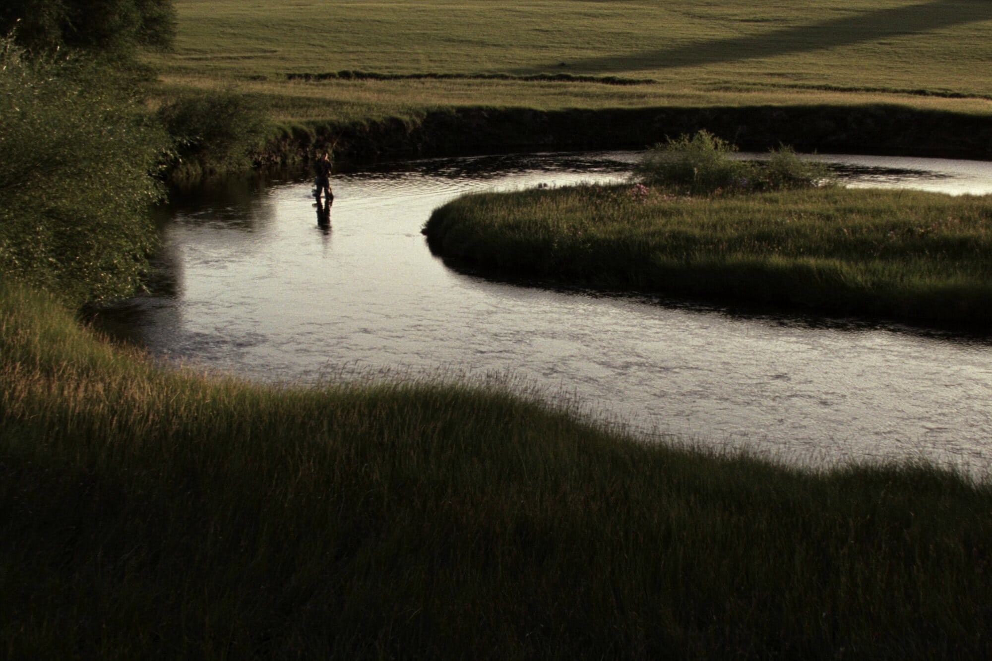 A lone angler casting on a private spring creek winding through a meadow in the late evening.