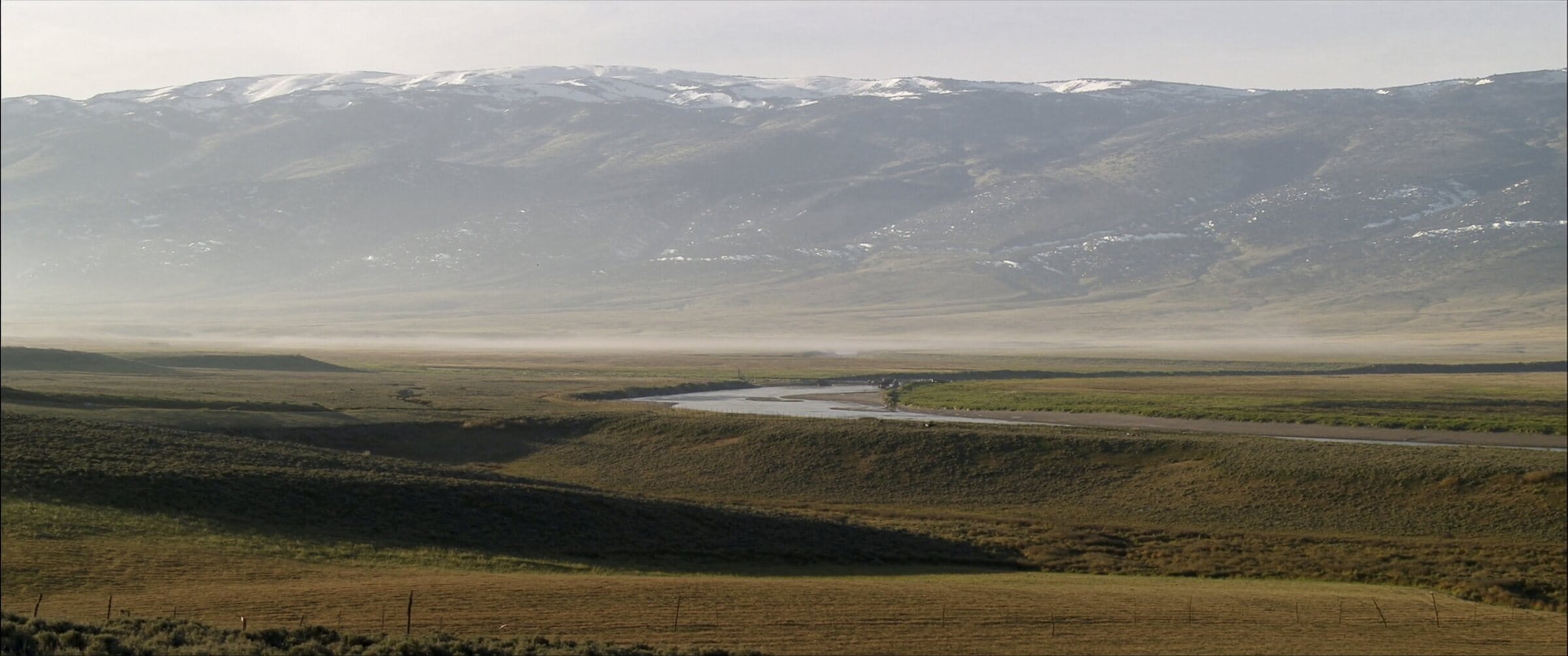 A wide Western ranch valley at first light, a river glinting through hayfields below snow-dusted peaks.